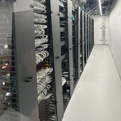 A row of server cabinets with cables arranged in a data center hallway, under bright fluorescent lighting — showing the construction of a professional server farm by Tal Systems.