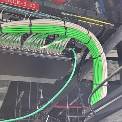 Green and white cable braids are neatly connected and connected to a board mounted on a rack and marked "RACK-3-03" on the ceiling of Tal Systems' corporate server farms.