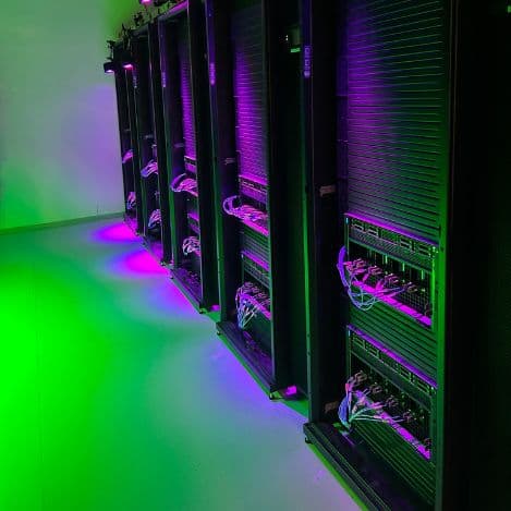 Rows of server cabinets in a data center are lit with green and purple lights, with network cables connected to the equipment, showcasing Tal Systems and Server Farm Maintenance professionals at work.