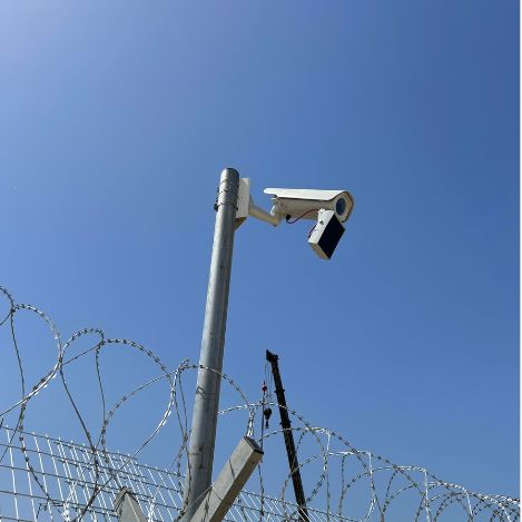 A security camera mounted on a pole overlooks a barbed wire fence under clear blue skies, securing the server farm for companies.
