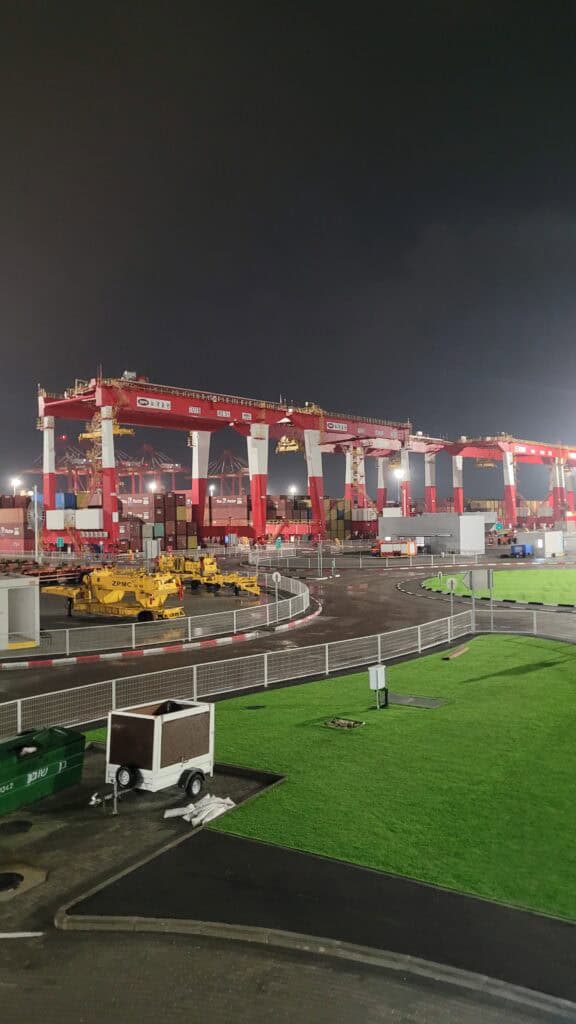 Night view of a container port with large red cranes, shipping containers and industrial vehicles under artificial lighting.